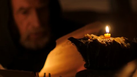 Kiev/ukraine - Jul 14 2015: Dark Room in an Old Log Hut of the 11Th Century. Candlelight. Old Man, Monk, Chronicler, Dressed in Black Robe, in the Hood. Nestor the Chronicler. he Writes With a Quill
