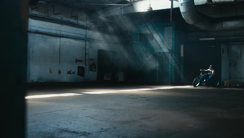 Young Caucasian male in denim overalls with tattoos riding his custom made bicycle in large warehouse garage. 60 FPS slow motion Blackmagic URSA Mini RAW graded footage