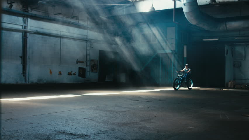 Young Caucasian male in denim overalls with tattoos riding his custom made bicycle in large warehouse garage. 60 FPS slow motion Blackmagic URSA Mini RAW graded footage