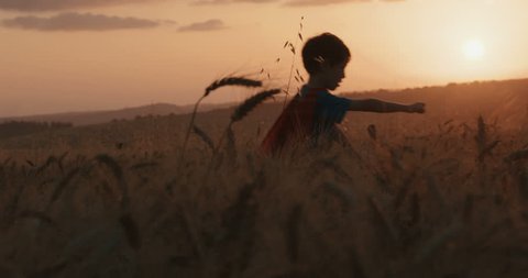 Boy with a super hero cape runs in a field during sunset
