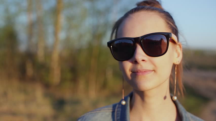 Young, stylish, informal girl looks around, takes off sunglasses, turns toward the sun, squinting from the sun, dresses sunglasses, middle distance.