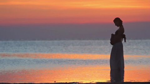 Silhouette of pregnant woman at the beach at sunset. Pregnant lady standing at the sea and caress her belly.