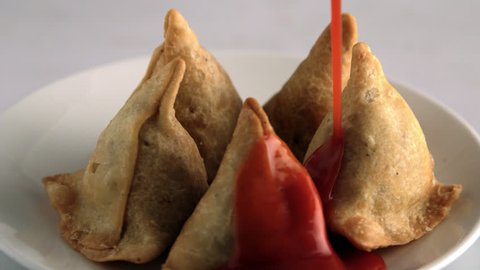 This is a shot of Samosas (samosa) revolving , as ketchup is pouring on. Delicious samosas with Red color tomato sauce pouring. Samosas are distinctly triangular. Shot at high speed on Phantom camera.