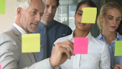Multi ethnic young businesswoman sticking adhesive notes on glass wall in office and boss circle post it. Close up of leadership and his business team looking at sticky notes on window in slow motion.