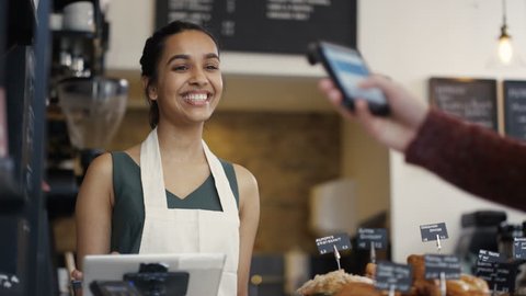 4K Cheerful worker serving a customer who uses smartphone to pay in coffee shop. UK - April, 2016