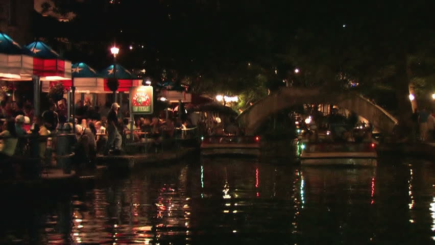 San Antonio Texas Riverwalk. Tourists at restaurants and shops. Vacation area. Tourists eating at outside tables with bright lights and festive atmosphere. Tour boat crosses by. 