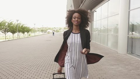 Full-length side view of slim african american business lady in short dress and high boots going to the airport with her luggage. Business trip concept