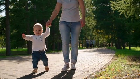 Adorable baby boy walking in the autumn park with his mom holding hands in slow motion