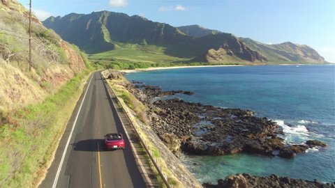AERIAL: Red convertible car driving along the coastal road above dramatic rocky shore towards volcanic mountains. Happy young couple on summer vacation traveling at the seaside in Oahu island, Hawaii