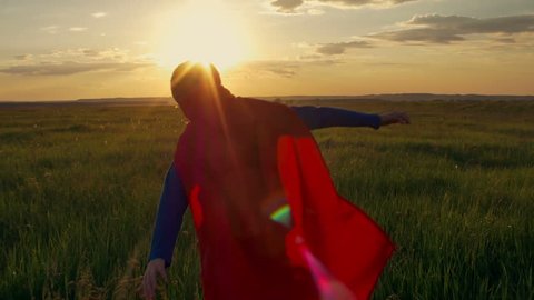Boy dressed with a Superman cape running in a field, looking into the sunset. A boy dressed as a superhero runs through a wheat field. Children's games, child's dream. Child playing in field at sunset