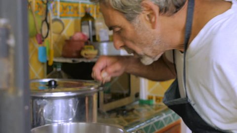 A man in his 60s tastes soup out of a large pot and adds salt without measuring the amount.