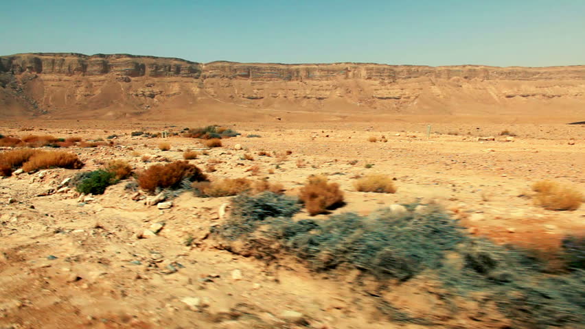 Wide shot drive-by of the sparsely foliaged desert floor and the far off cliff