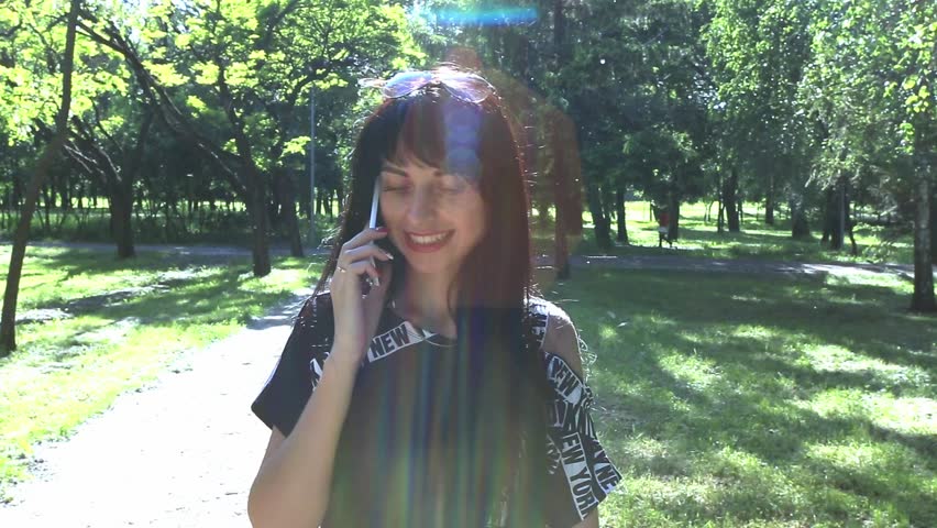 Beautiful brunette girl walking in park and speaks by phone at summer day with backlight