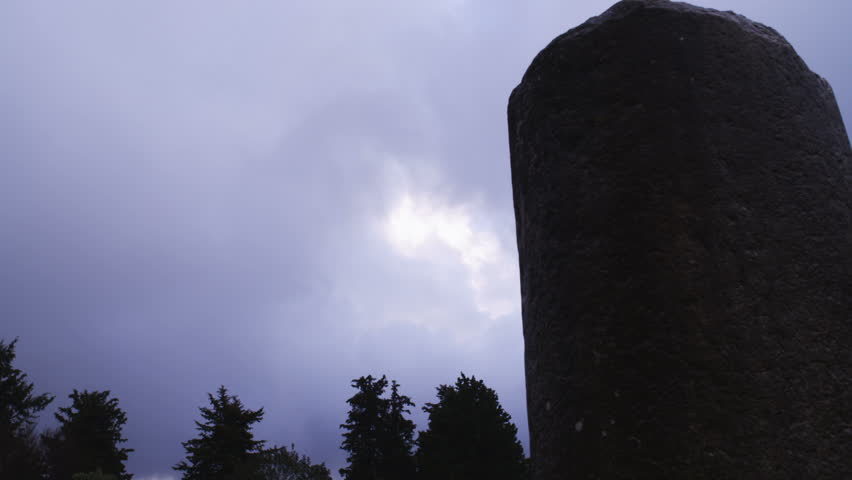 A shot of the sun through the heavy clouds, past a silhouette column and the tops of some pine trees, with a small tilt down at the end of the shot at the synagogue of Bar