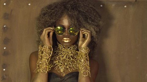 Magic surreal golden african american female model in massive sunglasses with bright glitter makeup, glossy golden hairstyle and big red lips