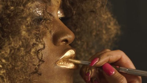 Close-up of magic golden african american model with bright glitter makeup and golden glossy hairstyle preparing for shooting at the stylist in studio backstage