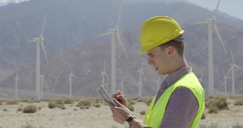 Technician in hard hat and wearing yellow high-visibility vest, at wind farm, using a tablet computer and stylus. Walks off frame at end. Medium close up, originally recorded in 4K.