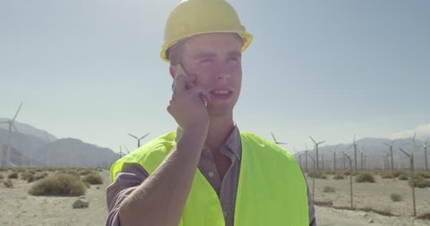 Technician in hard hat and wearing yellow high-visibility vest, at wind farm, taking a phone call as camera dollies around him. Close up, originally recorded in 4K.
