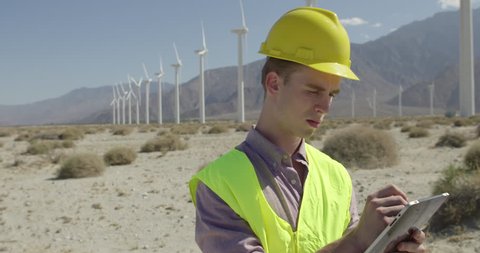 Dolly around wind farm technician wearing hard hat and safety vest, using a tablet computer and stylus. Medium close up, originally recorded in 4K slow motion at 60fps.