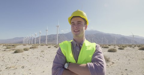 Wind farm technician in hard hat and wearing yellow high-visibility vest, looking confident and happy as camera dollies around him. Medium shot, originally recorded in 4K.