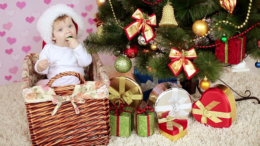 Babe in santa hat sitting in a basket near the Christmas tree and gifts