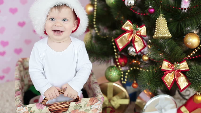 Laughing baby in santa hat standing in a basket near the Christmas tree and