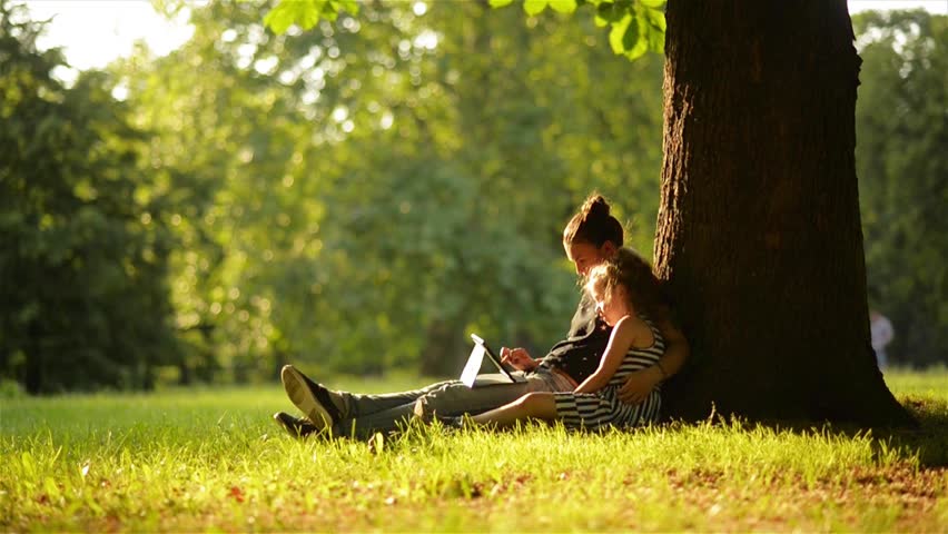 Young mother and her daughter playing on a tablet and a dog in a beautiful park
