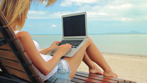 Young woman sitting at the sunbed with a laptop in front of sunset view. Lady freelancer working at the beach.
