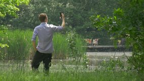Young Man Waves His Hand Into Mobile Phone in Green Park, Making Videocall Near the Lake, Taking Selfie, Friendly Man With Smartphone Clicks the Screen, Man in Glasses Turns and Walks Away, Jumps