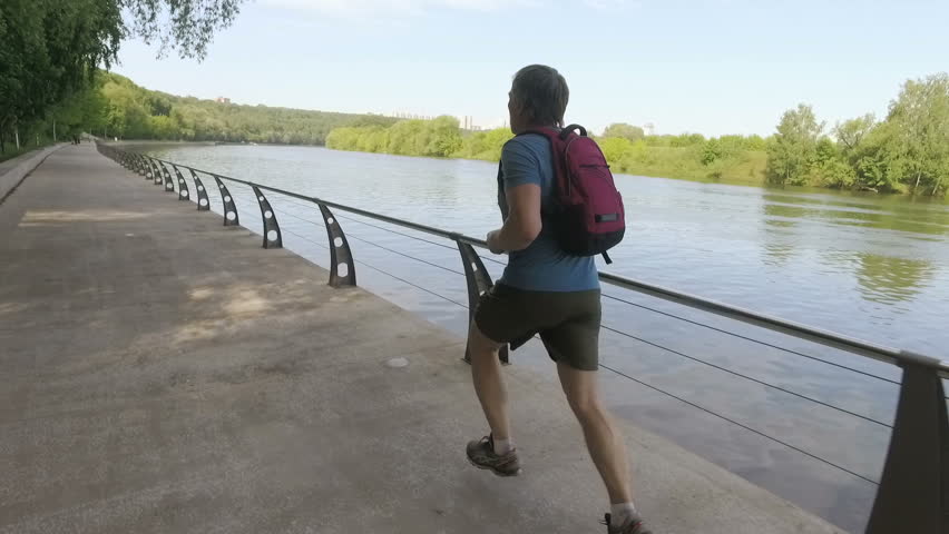 Adult man with backpack training on embankment against river in the morning in slow motion