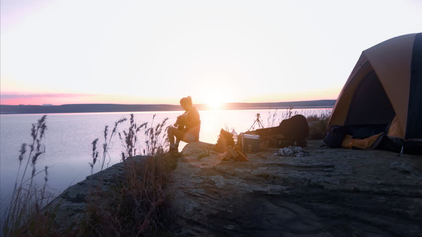 wide shot of young alternative independent woman playing guitar next to the fire and tent on camping trip at serene beautiful lake
