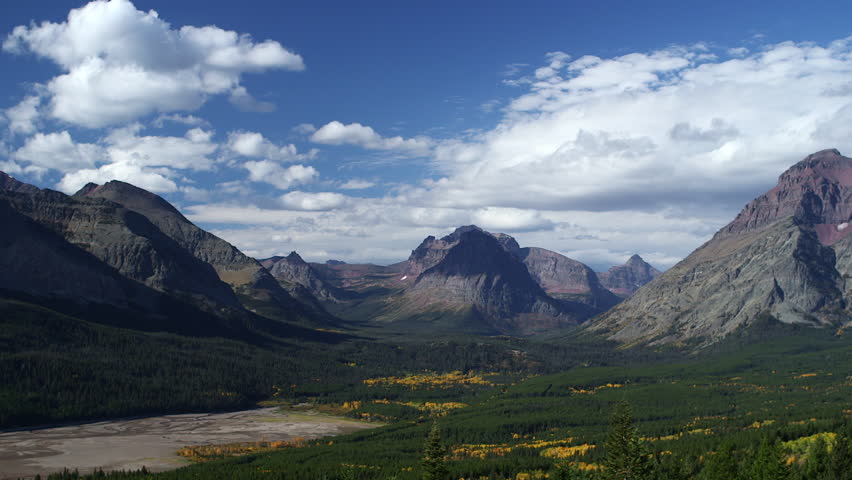 Jagged wild granite peaks frame a vast glacial valley as white clouds cruise by. There are yellow aspen trees visible in the valley, as well as a dry lake drainage: Glacier, Montana - September, 2015