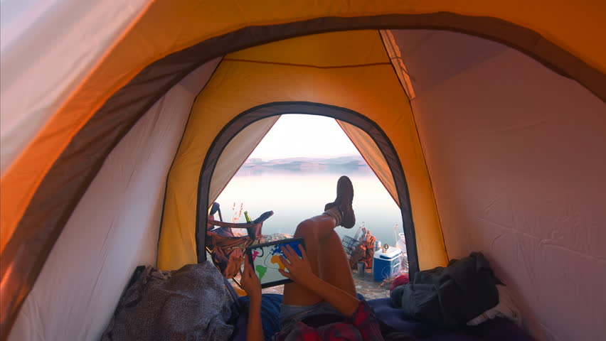 4k Medium shot of epic young traveler woman using a tablet to view a world map whilst chilling in her tent on camping adventure.
