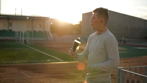 The tired athlete with naseball cap drinks water from the bottle on stadium track