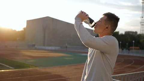 The tired athlete with naseball cap drinks water from the bottle on stadium track