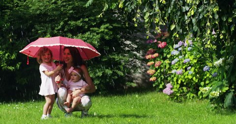 little girl in the garden playing with parents hiding under a red umbrella against splashing water and looking in camera . concept of security and saving of energy 