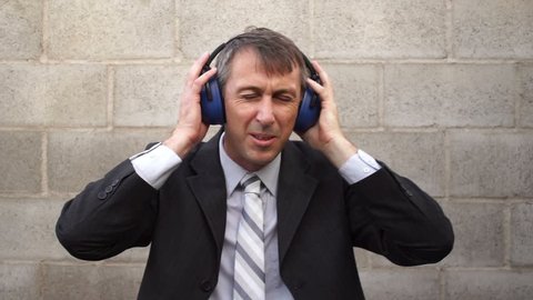 Man wearing a business suit and tie against a cinder-block wall with noise blocking safety acoustic earmuffs  grimacing in pain from loud noises.