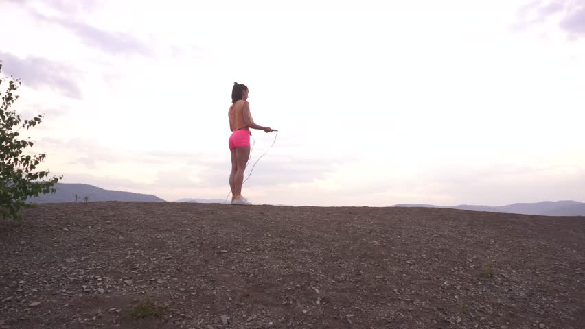 Back view on active young brunette woman jumping with skipping rope on the mountain peak. Green mountain ladscape background. Outdoor training concept