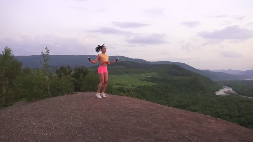 Active young brunette woman jumping with skipping rope on the mountain peak. Green mountain ladscape background