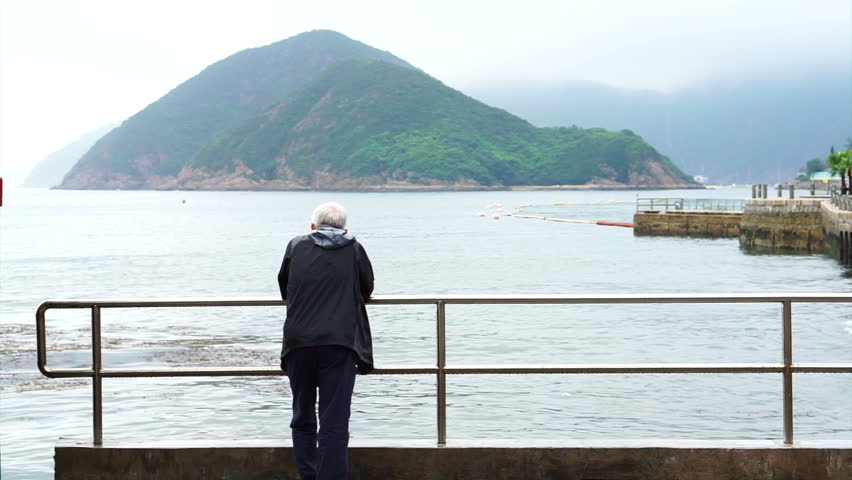 Asian senior man standing alone at the beach front pier in gloomy rainy day