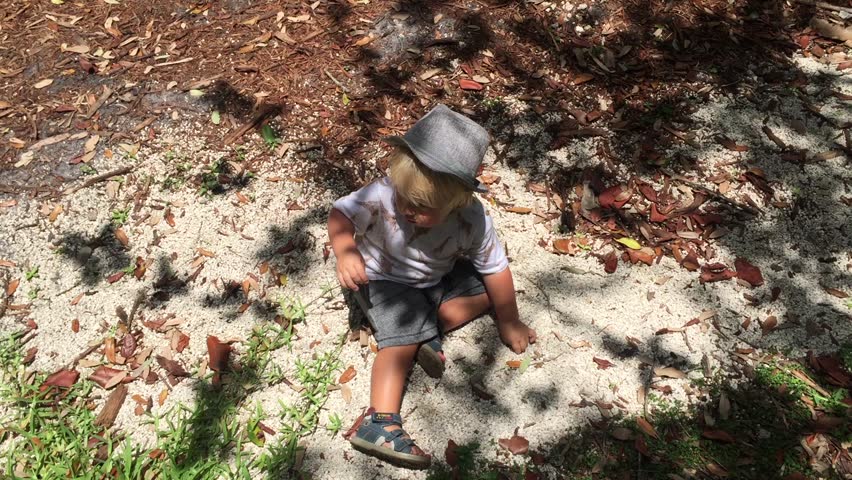  A blonde boy in a hat is sitting on the sand. There