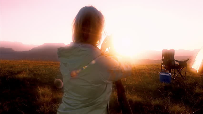 Female traveler and photographer taking pictures of the beautiful sunset after a hike with red backpack next to yellow tent. 4k. scenic.natural
