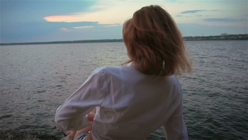 Tender girl in white blouse looking at the river at sunset. Slow motion