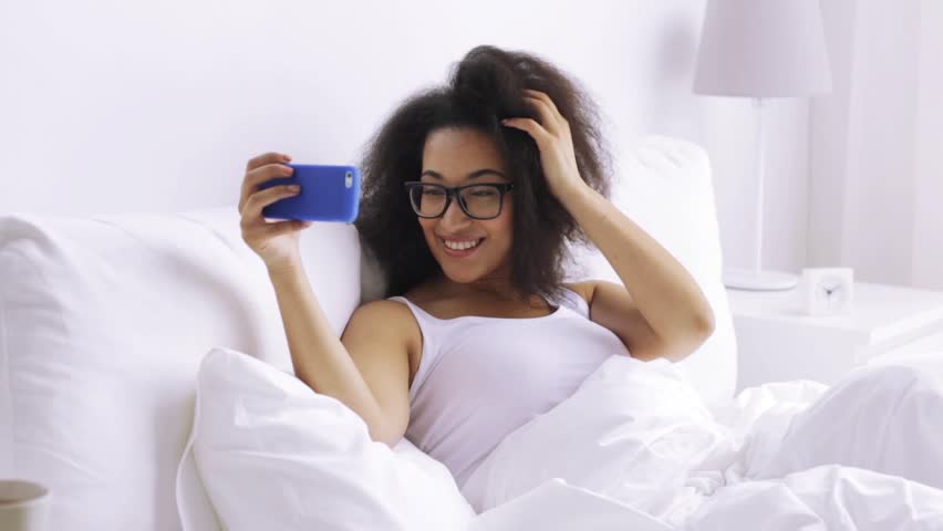 rest, technology and people concept - young african woman smartphone taking selfie in bed at home bedroom