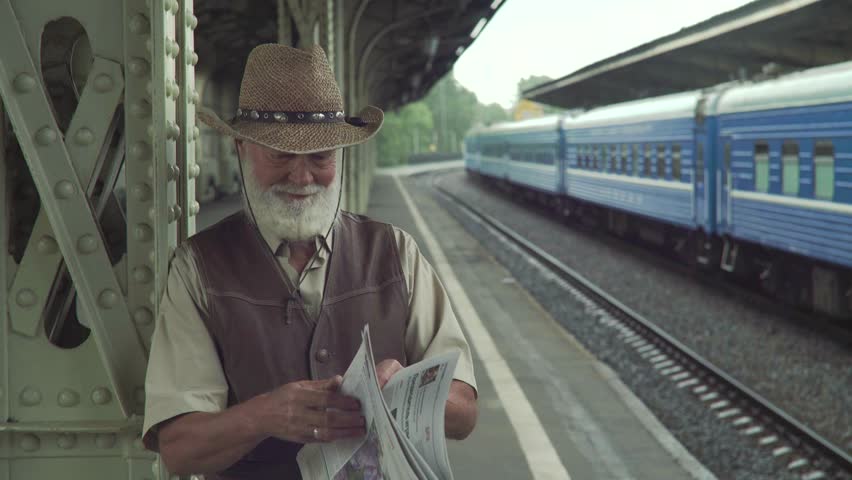 Senior gentleman with hat reading a newspaper on the platform of train station