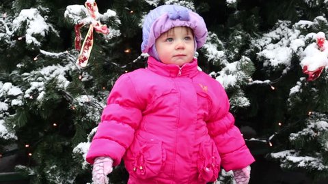 Little girl stands and look toward in front of Christmas tree covered with snow, and then she raises her arms up