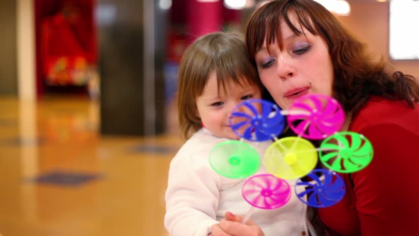 Daughter sit on mothers knees, they blow in fan colorful toy