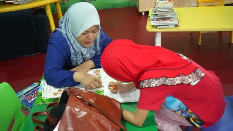 Asian Muslim mom wearing headscarf helping kids reading books, and finishing homework in a children library corner.