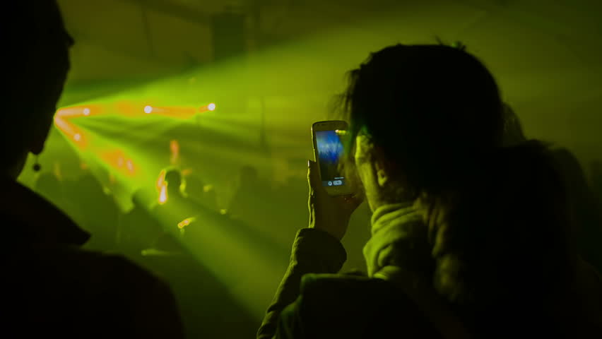 Some people are dancing at a rock concert and the other people are recording different songs. The light show is going on, too. Close-up shot.