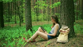 Beautiful girl in the woods for a walk, sitting under a tree and read Tablet PC. Video is accompanied by the sounds of birds singing in the forest. Outdoor hipster girl with tablet